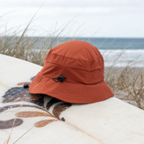Burnt Orange surf hat with bump protection and a elastic toggle adjustment on a surfboard with a beach background