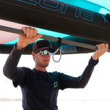 A man holds the boom of a wing foil wing branded with 'OZONE' in blue lettering, showing a bungee harness line attached to the boom.