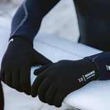 Close-up of a person wearing black Nuwave 3.0 gloves while sitting on a white surfboard outdoors.