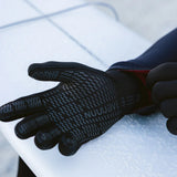 A close-up of a black wetsuit glove with a textured grip pattern and the brand name 'NUUJUAVE' printed on the back, worn on a hand resting on a white surfboard.