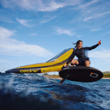 A person in a black wetsuit riding a black wing foil board on a wave while flagging a yellow and grey wingfoil wing with the branding 'OZONE' and 'ALLULA' on the leading edge