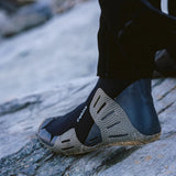 A close-up of a person's foot wearing a black and gray wetsuit boot on a rocky, outdoor surface. with white branding on side saying ' C-SKINS'