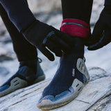 A person wearing a black wetsuit and gloves is adjusting a black Neoprene bootie on a rocky beach. The bootie has a red cuff and gray accents, with the brand name 'c-skins' visible on the side.