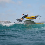 A foiler at the top of a wave with the foil out the water on a turn holding a wingfoil wing in colours grey and yellow flagginf balanced behind foiler on the wave