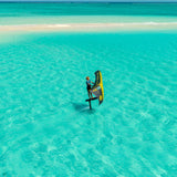 Aerial view of a person wingfoiling with a grey and yellow branded OZONE wing in ALLULA Fabric on clear turquoise ocean water near a narrow sandy shoreline under a blue sky.
